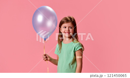 Girl with long hair wearing green shirt, and medicine patch on hand, holding pastel blue balloon with happy smile against pink background. 121450065