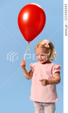 Little charming child, girl in pink clothes, smiling softly while holding red balloon against blue studio background. 121450345