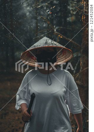 Warrior woman in a conical hat and black mask walks in a mystical forest on a rainy day, holding a katana sword. The scene blends fantasy and japanese culture, with lush nature and towering trees. Warrior woman in a conical hat and black mask walks in a mystical forest on a rainy day, holding a katana sword. The scene blends fantasy and japanese culture, with lush nature and towering trees. 121452084