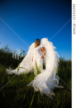 A blonde with large white angel wings stands in a field of tall grass under a clear blue sky, creating a serene and ethereal scene. Cute angel against a blue sky. 121452085