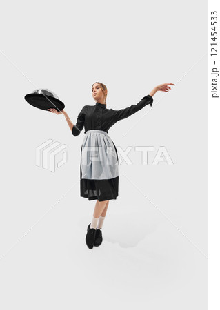 Woman holding crystal glass presents elegance, dressed in black attire with apron against white studio background. Wide angle photo. 121454533