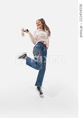 Energetic woman with paint roller jumps enthusiastically in denim pants splattered with paint, highlight style against white studio background. Energetic woman with paint roller jumps enthusiastically in denim pants splattered with paint, highlight style against white studio background. 121454556