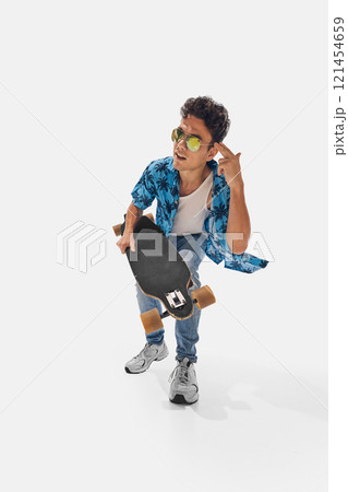 Skateboarder in blue palm-printed shirt and ripped jeans crouching playfully, gesturing with skateboard in hand. Wide angle photo. 121454659