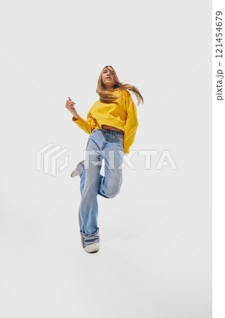 Woman wearing yellow sweater and blue jeans energetically jumping with loose hair flowing freely against white studio background. 121454679