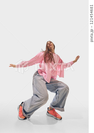 Woman squats slightly, while dancing dressed in casual pink and denim, with expressive confidence against white studio background. 121454681