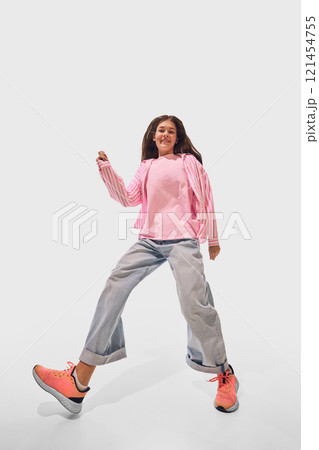 Wide angle photo of woman in pink striped shirt and loose denim playfully dancing expressing movement and balance against white studio background. 121454755