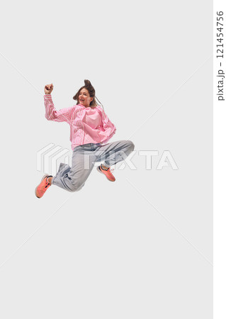 Woman in casual pink and denim leaps mid-air with joyful expression, showcasing energy and motion against white studio background. 121454756