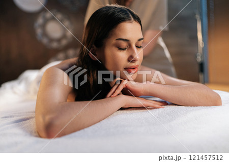 Closeup face of calm woman client relaxing during back massage in spa salon with dark interior. Professional massage therapist providing therapeutic back massage to relaxed female, close-up. Closeup face of calm woman client relaxing during back massage in spa salon with dark interior. Professional massage therapist providing therapeutic back massage to relaxed female, close-up. 121457512