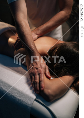 Vertical cropped shot of massage therapist performing deep tissue technique on female shoulder, targeting muscle tension and promoting wellness during bodywork session in spa salon with dark interior Vertical cropped shot of massage therapist performing deep tissue technique on female shoulder, targeting muscle tension and promoting wellness during bodywork session in spa salon with dark interior 121457517