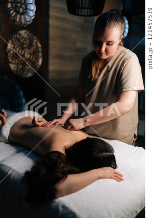 Vertical shot of focused masseuse giving relaxing back massage to relaxed young woman lying on massage table in spa, creating tranquil and rejuvenating experience. Concept of professional massage. 121457519