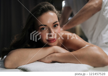 Close-up portrait of beautiful young woman looking at camera enjoying moment of wellness and pampering, getting massage in beauty parlor. Masseuse giving relaxing back massage to female client. Close-up portrait of beautiful young woman looking at camera enjoying moment of wellness and pampering, getting massage in beauty parlor. Masseuse giving relaxing back massage to female client. 121457520