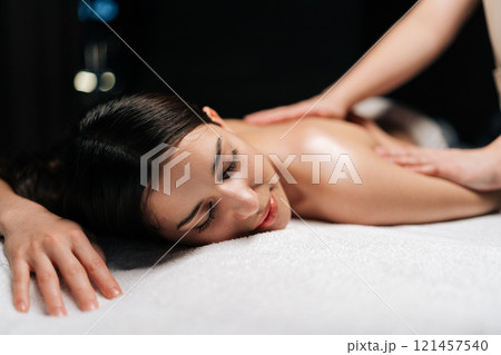 Close-up portrait of beautiful calm young woman client relaxing during back massage in spa salon with dark interior. Professional massage therapist providing therapeutic back massage to relaxed female Close-up portrait of beautiful calm young woman client relaxing during back massage in spa salon with dark interior. Professional massage therapist providing therapeutic back massage to relaxed female 121457540