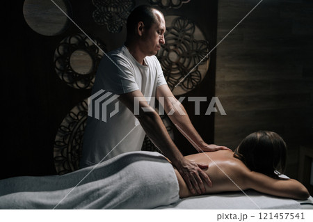 Side view of professional male massage therapist providing therapeutic back treatment, working female client lying on table in wellness center with dark interior. Concept of rest with spa treatment. Side view of professional male massage therapist providing therapeutic back treatment, working female client lying on table in wellness center with dark interior. Concept of rest with spa treatment. 121457541