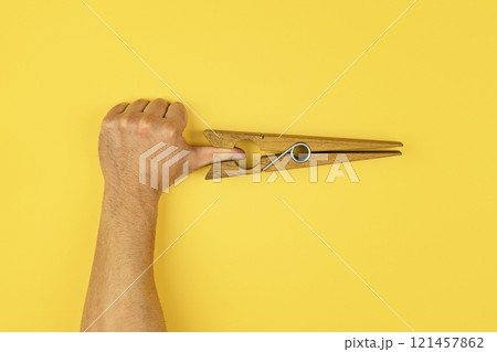 Creative composition of human hand gripped by large wooden clothespin, placed against vibrant yellow background, perfect for quirky visual concepts 121457862