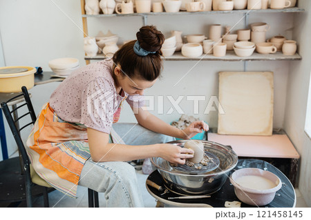 Young woman sculpting pottery on the wheel in a bright studio space Young woman sculpting pottery on the wheel in a bright studio space 121458145
