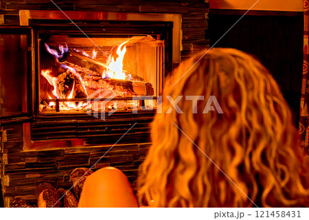 Blonde curly haired woman sitting in front of fireplace fire warming herself from cold winter. Logs of firewood Blonde curly haired woman sitting in front of fireplace fire warming herself from cold winter. Logs of firewood 121458431