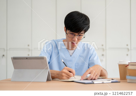 A smart Asian male medical student in scrubs, focused on studying at his desk in a classroom. A smart Asian male medical student in scrubs, focused on studying at his desk in a classroom. 121462257