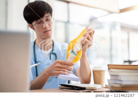 A smart Asian male medical student in scrubs is examining a model of bone joints. 121462287