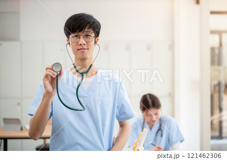 A smart Asian male medical student in scrubs, smiling and showing his stethoscope to the camera. 121462306