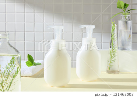 In the center are two white pump bottles no label, surrounded by some glass vases containing a green tea leaves and fern branches, place on a yellow surface, in a bathroom backdrop. Advertising photo. In the center are two white pump bottles no label, surrounded by some glass vases containing a green tea leaves and fern branches, place on a yellow surface, in a bathroom backdrop. Advertising photo. 121468008