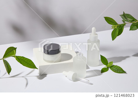 Mockup of a jar and serum bottles no label, set on a white pedestal with green tea leaves - anti-acne and moisturizing benefits. Side view. A photo to promote skincare products 121468023
