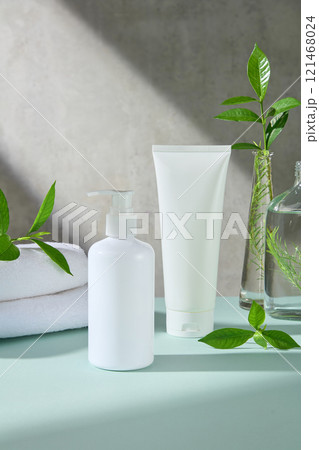 A close-up of a set of unlabeled products, a white tube and a white pump bottle, placed on a blue surface, next to glass vases containing green tea leaves and tea tree branches, in a grey backdrop. 121468024