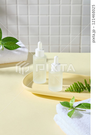 Mockup of dropper bottles placed on a wooden tray, next to some towels, green tea leaves, and fern fronds, in a bathroom setting. Advertising photos for serum products. 121468025