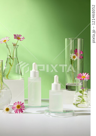Close-up of an unlabeled mockup of dropper bottles on white and glass podiums, surrounded by some lab glassware with chamomile flower branches, a green backdrop for a cosmetics advertisement photo. Close-up of an unlabeled mockup of dropper bottles on white and glass podiums, surrounded by some lab glassware with chamomile flower branches, a green backdrop for a cosmetics advertisement photo. 121468052