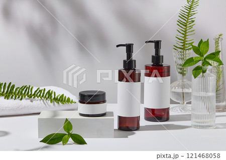 A frontal view of a skincare product ad features a mockup of a jar and two pump bottles set on a white platform. Surrounded by green tea branches, tea plants, and ferns in glass vases and towels. A frontal view of a skincare product ad features a mockup of a jar and two pump bottles set on a white platform. Surrounded by green tea branches, tea plants, and ferns in glass vases and towels. 121468058