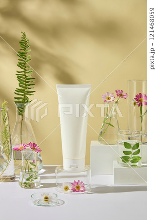 Close-up view of an unbranded cosmetics tube mockup, displayed on a glass podium, decorated by fresh tea trees, ferns, and chamomile branches. Mockup for vegan cosmetics advertisement photo. 121468059
