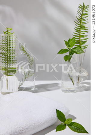 Spa concept with a few glass vases filled with some fresh green teas, ferns, and tea tree leaves, next to empty towels for display products, against a light background. 121468072
