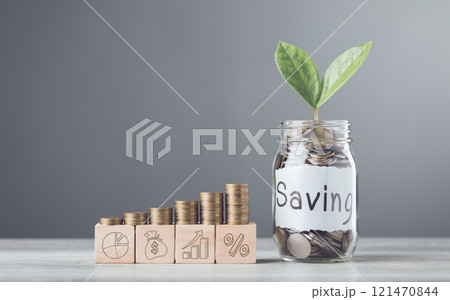 A glass jar filled with coins and a sprout labeled Saving, wooden blocks marked 2025, and neatly stacked coins on a table, symbolizing financial growth and future aspirations against a gray backdrop A glass jar filled with coins and a sprout labeled Saving, wooden blocks marked 2025, and neatly stacked coins on a table, symbolizing financial growth and future aspirations against a gray backdrop 121470844