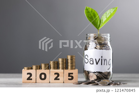 A glass jar filled with coins and a sprout labeled Saving, wooden blocks marked 2025, and neatly stacked coins on a table, symbolizing financial growth and future aspirations against a gray backdrop A glass jar filled with coins and a sprout labeled Saving, wooden blocks marked 2025, and neatly stacked coins on a table, symbolizing financial growth and future aspirations against a gray backdrop 121470845