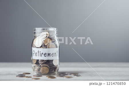 Glass jar filled with coins and a sprout labeled 'Retirement,' placed on a table with a gray background, symbolizing saving for financial growth and secure future planning 121470850