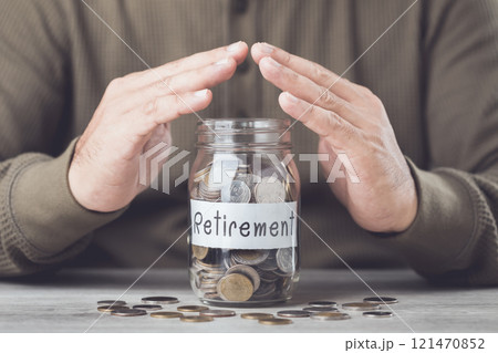 A glass jar filled with coins, labeled 'Retirement,' as a man uses both hands to form a protective symbol, representing the safeguarding of his financial future 121470852