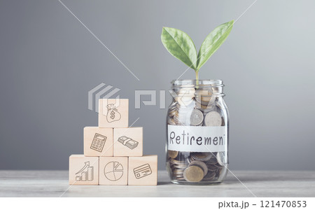 Glass jar with coins and a sprout labeled 'Retirement,' alongside stacked wooden blocks with financial symbols, placed on a table against a gray background, representing savings growth Glass jar with coins and a sprout labeled 'Retirement,' alongside stacked wooden blocks with financial symbols, placed on a table against a gray background, representing savings growth 121470853