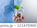 Glass jar filled with coins, with a small seedling growing in the center, placed on a table against a dark grey backdrop. The image represents the concept of saving and financial growth for future Glass jar filled with coins, with a small seedling growing in the center, placed on a table against a dark grey backdrop. The image represents the concept of saving and financial growth for future 121470900