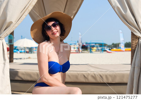 Woman in blue swimsuit sits in shade of beach cabana bungalow under canopy Woman in blue swimsuit sits in shade of beach cabana bungalow under canopy 121475277