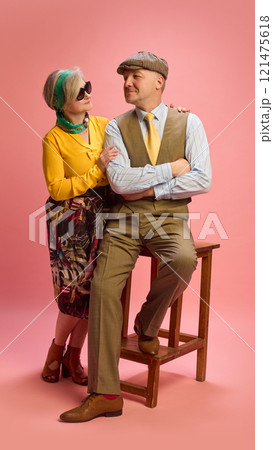 Woman in yellow blouse and green scarf leans on man's shoulder while he sits in brown trousers and yellow tie, both posing calmly against pink studio background. 121475618