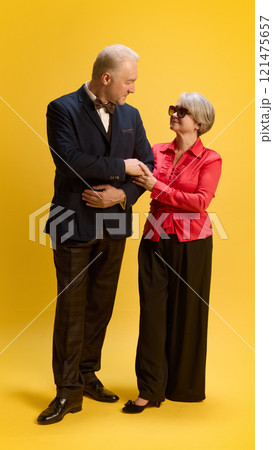 Couple exchanging smiles, man in formal wear leaning slightly toward woman in elegant outfit, standing with arms crossed, against yellow studio background. 121475657