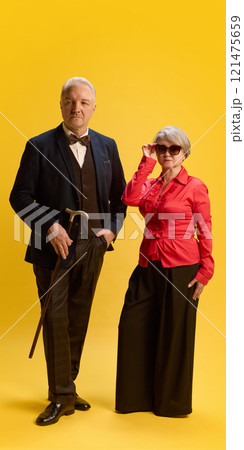 Mature couple, man in plaid blazer and scarf with woman in cream jacket and sunglasses, posing arm-in-arm against blue studio background. 121475659