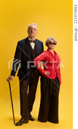 Elderly man in bow tie and formal suit holding cane, standing confidently beside woman in ruffled red blouse and black trousers, against yellow studio background. Elderly man in bow tie and formal suit holding cane, standing confidently beside woman in ruffled red blouse and black trousers, against yellow studio background. 121475661