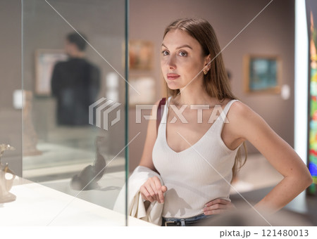 Young female visitor stands near glass display case and examines valuable exhibit Young female visitor stands near glass display case and examines valuable exhibit 121480013
