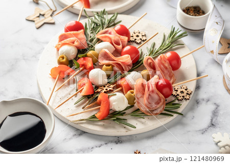Snack skewers with mozzarella, ham, olives, peppers, tomatoes. Festive canapes or antipasti on white tray for new year table Snack skewers with mozzarella, ham, olives, peppers, tomatoes. Festive canapes or antipasti on white tray for new year table 121480969