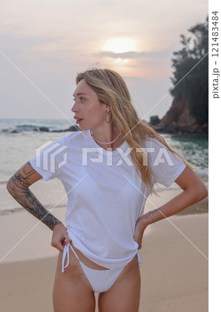A stylish woman in a white tee shirt enjoys a vibrant sunset at the beach, soaking in the moment 121483484