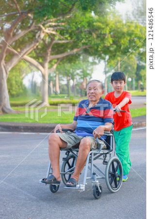 Grandson is happily pushing his grandfather's wheelchair in the garden. Boy takes care of the elderly. Senior man in wheelchair and with a grandchild. Concept of Family, Elderly, Therapy, Medical. 121486546