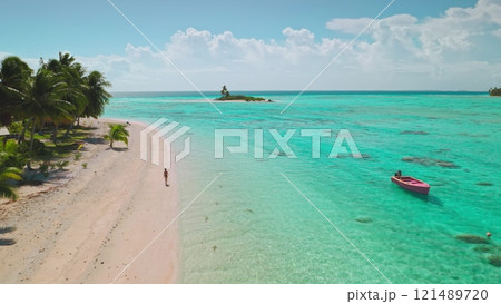 Aerial view of a tourist walking along a beautiful pink sand beach on a tropical island in aitutaki, cook islands, with a small boat anchored in turquoise water 121489720