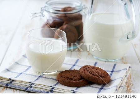 Glass and a jug of fresh milk with delicious chocolate brownie cookies on a white table, selective focus. Glass and a jug of fresh milk with delicious chocolate brownie cookies on a white table, selective focus. 121491140