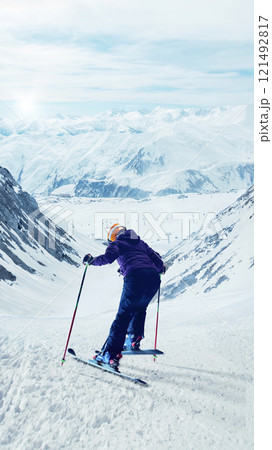 Skier standing on snow-covered slope, looking downhill at valley surrounded by snowy mountains and ski lifts in distance. 121492817