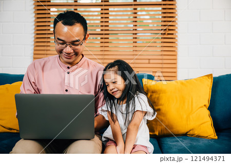 In a modern living room an Asian father combines work and family as he works on his laptop while his daughter learns on a computer for e-learning. Their togetherness and happiness shine through. 121494371
